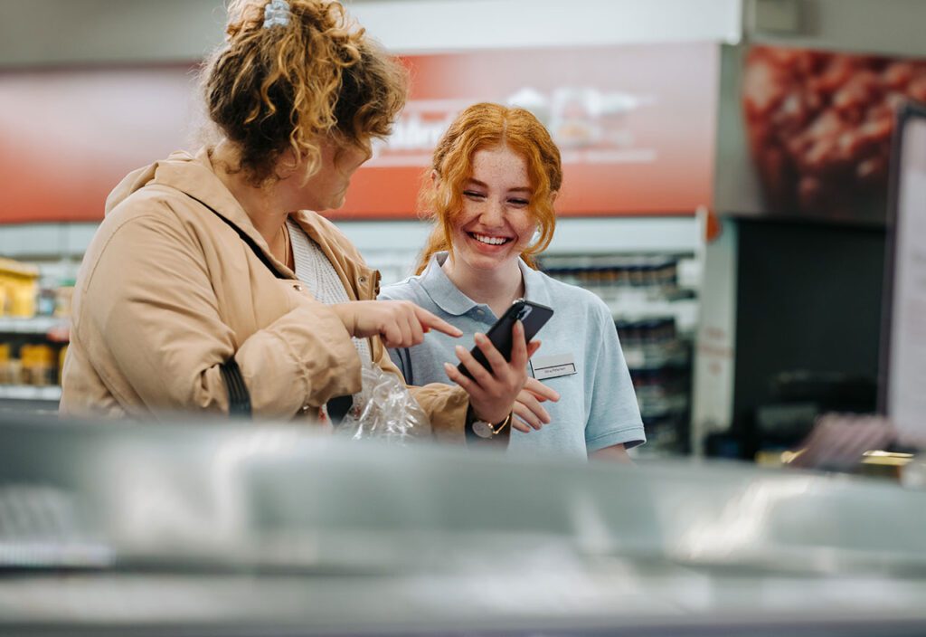 Smiling female employee at CPG store in front of POS while customer points to cell phone.